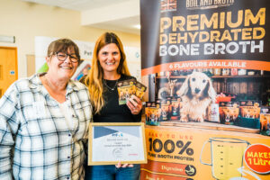 Boil & Broth Founder at the 2024 Business of Pets Conference holding her Bone Broth Product in her hand in front of her branded promotional banner.